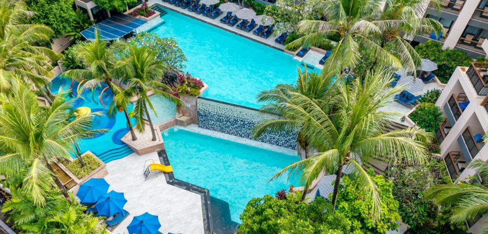 Aerial view of a resort's vibrant pool area, surrounded by lush greenery and palm trees. Blue sun umbrellas and lounge chairs add a relaxing, tropical vibe.