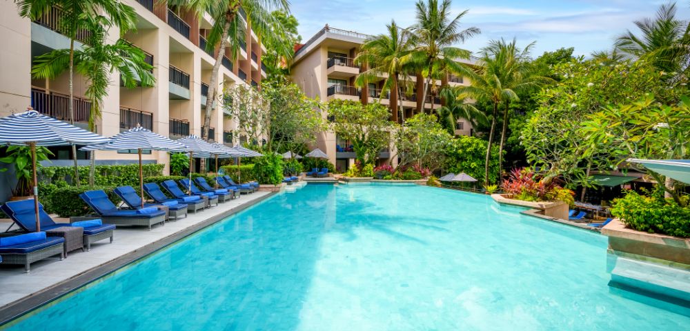 A serene hotel pool scene featuring clear blue water, surrounded by lush greenery and palm trees. Blue lounge chairs and striped umbrellas line the poolside.