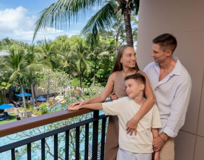 A family of three stands on a balcony overlooking a lush tropical garden and pool. The parents smile at each other while holding their son, conveying warmth and contentment.