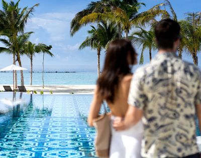 A couple stands by an infinity pool, facing the ocean. Palm trees frame the background under a clear blue sky, evoking a serene, tropical vibe.