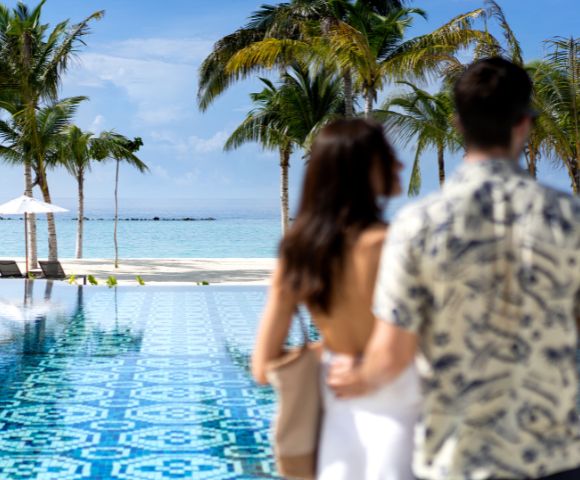 A couple in swimsuits relaxes on a blue outdoor bed, surrounded by a tropical beach setting with palm trees and a clear blue sea in the background.