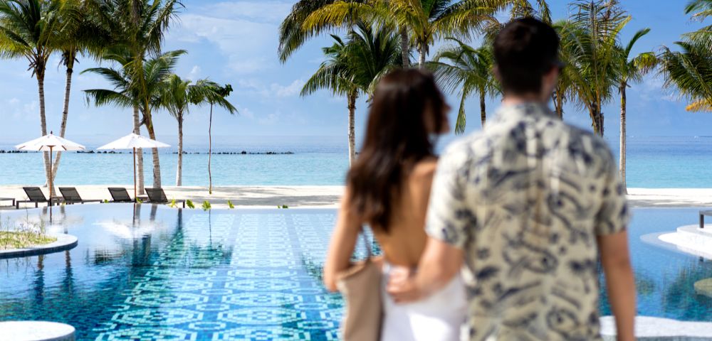 A couple in swimsuits relaxes on a blue outdoor bed, surrounded by a tropical beach setting with palm trees and a clear blue sea in the background.
