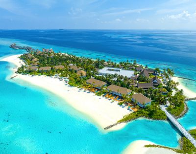 Aerial view of a tropical island resort with lush greenery, sandy beaches, and turquoise waters. Wooden beachfront bungalows and a pier create a tranquil scene.