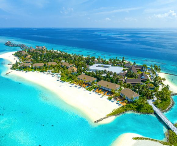 Aerial view of a tropical island resort with lush greenery, sandy beaches, and turquoise waters. Wooden beachfront bungalows and a pier create a tranquil scene.