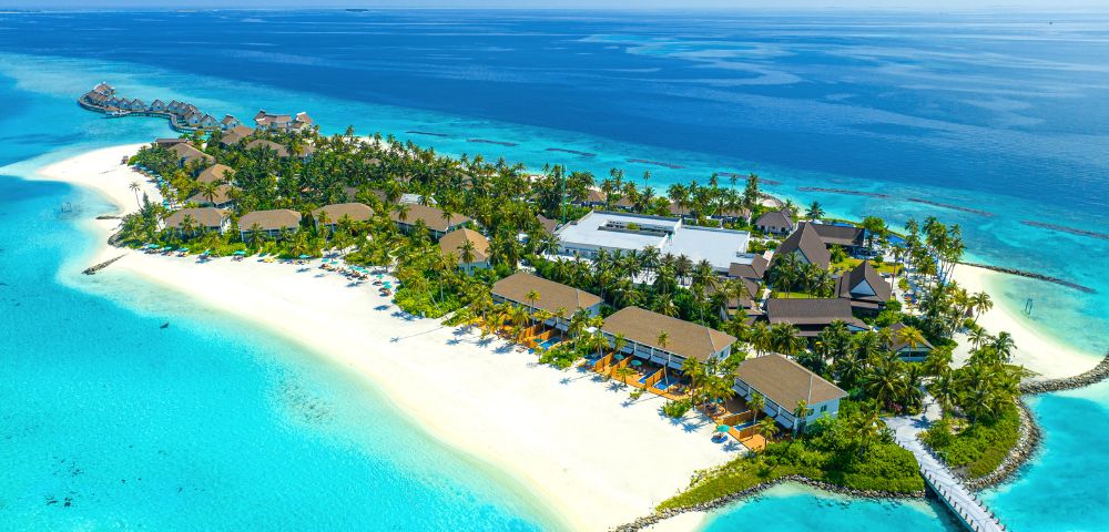 Aerial view of a tropical island resort with lush greenery, sandy beaches, and turquoise waters. Wooden beachfront bungalows and a pier create a tranquil scene.