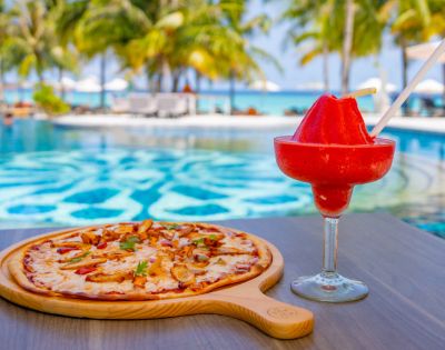 Pizza on a wooden board and a red frozen drink in a glass by a poolside, with palm trees and ocean in the background; a tropical and relaxed scene.