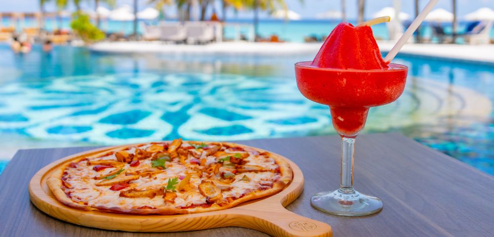 Pizza on a wooden board and a red frozen drink in a glass by a poolside, with palm trees and ocean in the background; a tropical and relaxed scene.