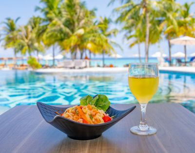 A bowl of pasta and a glass of orange juice sit on a table by a serene pool, with palm trees and a bright blue sky in the background, exuding a tropical vibe.