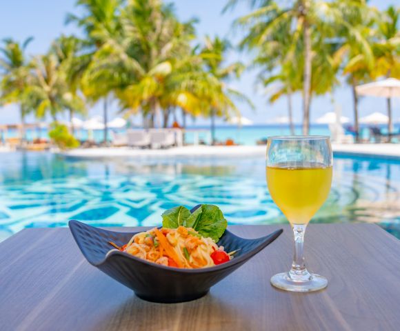 A bowl of pasta and a glass of orange juice sit on a table by a serene pool, with palm trees and a bright blue sky in the background, exuding a tropical vibe.