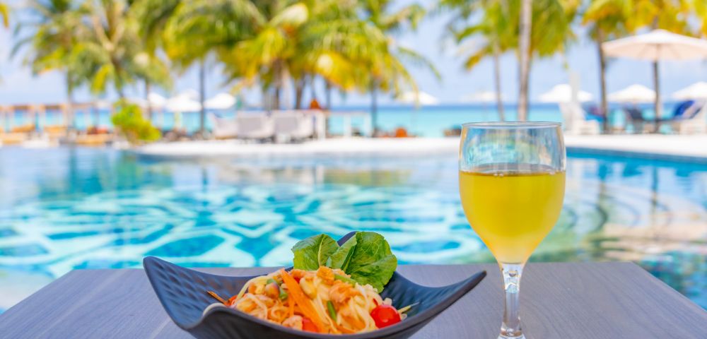 A bowl of pasta and a glass of orange juice sit on a table by a serene pool, with palm trees and a bright blue sky in the background, exuding a tropical vibe.