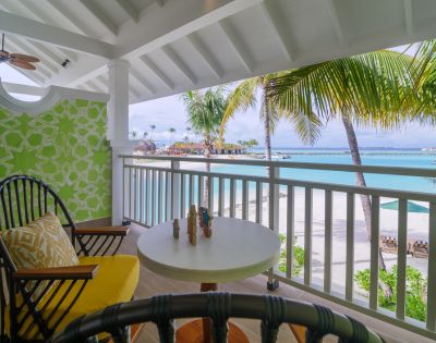 Covered balcony overlooking a beach with palm trees. Two chairs with yellow cushions surround a small table, conveying a tropical, relaxed atmosphere.