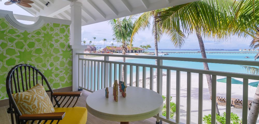 Covered balcony overlooking a beach with palm trees. Two chairs with yellow cushions surround a small table, conveying a tropical, relaxed atmosphere.