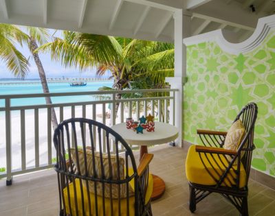 A tropical balcony with two wicker chairs and yellow cushions facing a table. Overlooks a beach with palm trees and turquoise water under a bright sky.