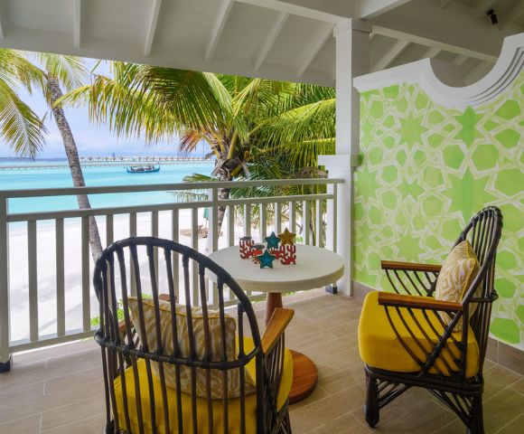 A tropical balcony with two wicker chairs and yellow cushions facing a table. Overlooks a beach with palm trees and turquoise water under a bright sky.