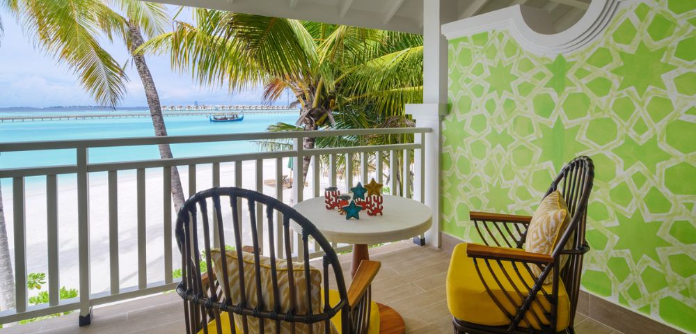 A tropical balcony with two wicker chairs and yellow cushions facing a table. Overlooks a beach with palm trees and turquoise water under a bright sky.