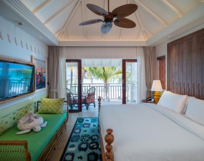 Bright, airy bedroom with a vaulted ceiling, ceiling fan, and wooden accents. A large bed and sofa face a balcony with tropical palm views. Calm and inviting.