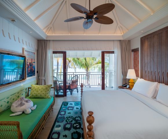 Bright, airy bedroom with a vaulted ceiling, ceiling fan, and wooden accents. A large bed and sofa face a balcony with tropical palm views. Calm and inviting.