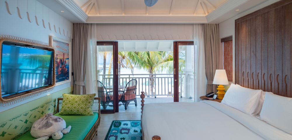 Bright, airy bedroom with a vaulted ceiling, ceiling fan, and wooden accents. A large bed and sofa face a balcony with tropical palm views. Calm and inviting.