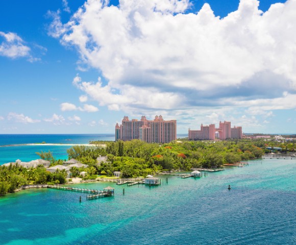 Aerial view of a tropical coastline with lush greenery and turquoise water. A pink luxury resort is visible in the background under a partly cloudy sky.