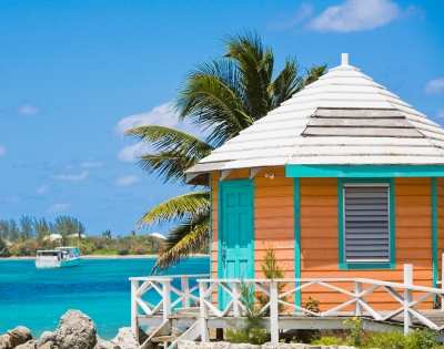 Tropical beach scene with a small, colorful beach hut featuring orange walls and turquoise accents. A palm tree leans over the hut, with blue ocean and a distant boat in the background under a clear sky. Peaceful and vibrant atmosphere.