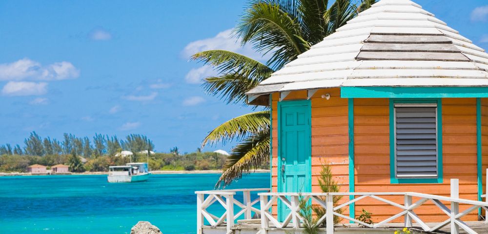Tropical beach scene with a small, colorful beach hut featuring orange walls and turquoise accents. A palm tree leans over the hut, with blue ocean and a distant boat in the background under a clear sky. Peaceful and vibrant atmosphere.