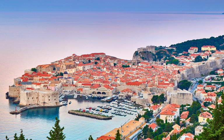 Panoramic view of Dubrovnik, Croatia. The historic city features orange-tiled roofs, stone walls, and a harbor with boats. Lush greenery surrounds the buildings.
