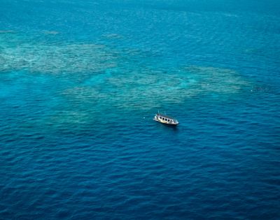 A lone boat floats on vibrant blue ocean water, surrounded by patches of visible sandy sea floor. The scene conveys tranquility and isolation.