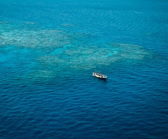 A lone boat floats on vibrant blue ocean water, surrounded by patches of visible sandy sea floor. The scene conveys tranquility and isolation.