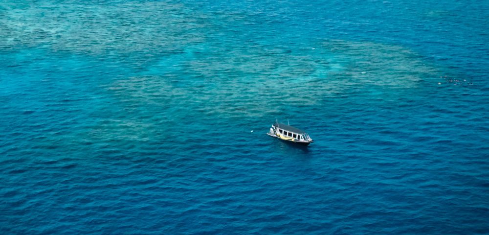 A lone boat floats on vibrant blue ocean water, surrounded by patches of visible sandy sea floor. The scene conveys tranquility and isolation.