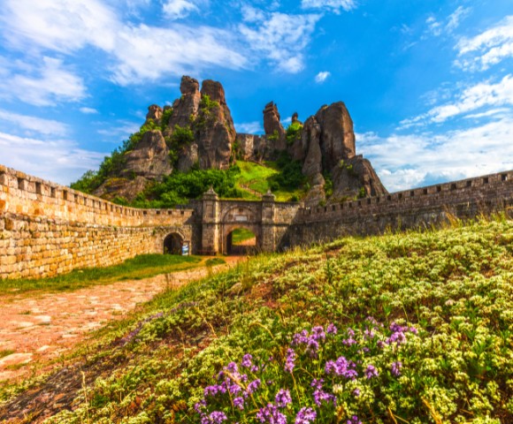Ancient stone fortress surrounded by lush greenery and wildflowers under a vibrant blue sky with scattered clouds. Majestic rock formations rise behind.