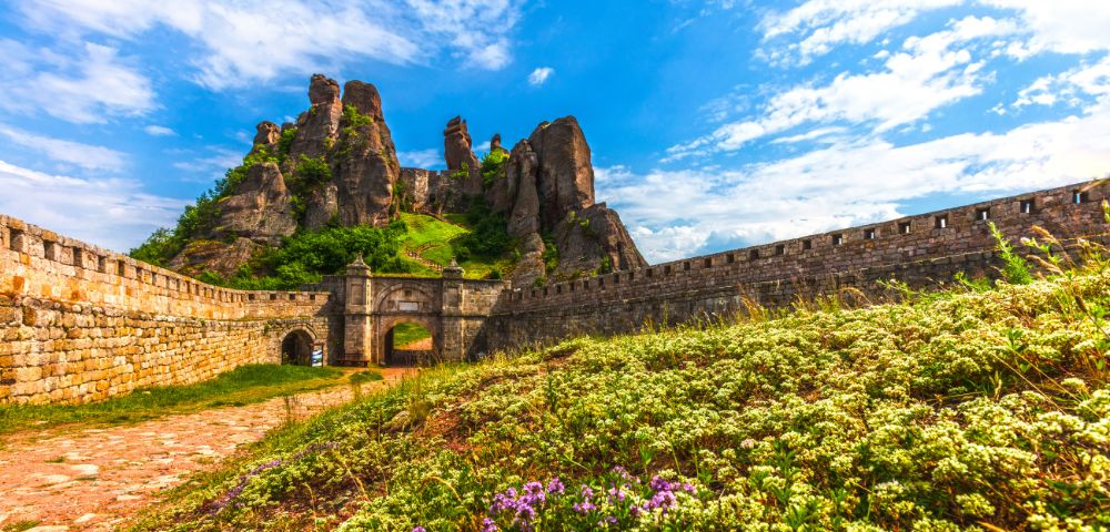 Ancient stone fortress surrounded by lush greenery and wildflowers under a vibrant blue sky with scattered clouds. Majestic rock formations rise behind.