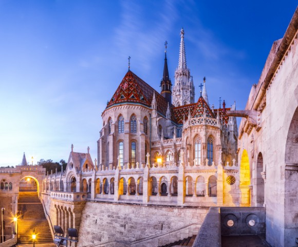 Gothic-style Fisherman's Bastion in Budapest at twilight, featuring ornate arches, a colorful tiled roof, and illuminated arches, creating a serene atmosphere.