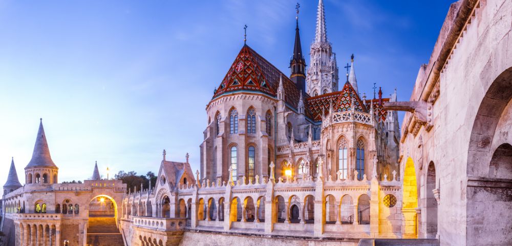 Gothic-style Fisherman's Bastion in Budapest at twilight, featuring ornate arches, a colorful tiled roof, and illuminated arches, creating a serene atmosphere.