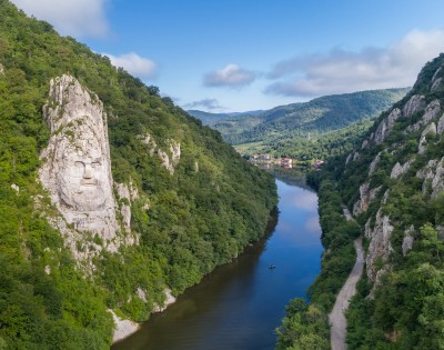 A serene river winds through a lush green canyon, flanked by steep hills. A large stone face is carved into one hill, under a clear blue sky.