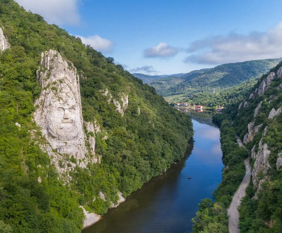 A serene river winds through a lush green canyon, flanked by steep hills. A large stone face is carved into one hill, under a clear blue sky.