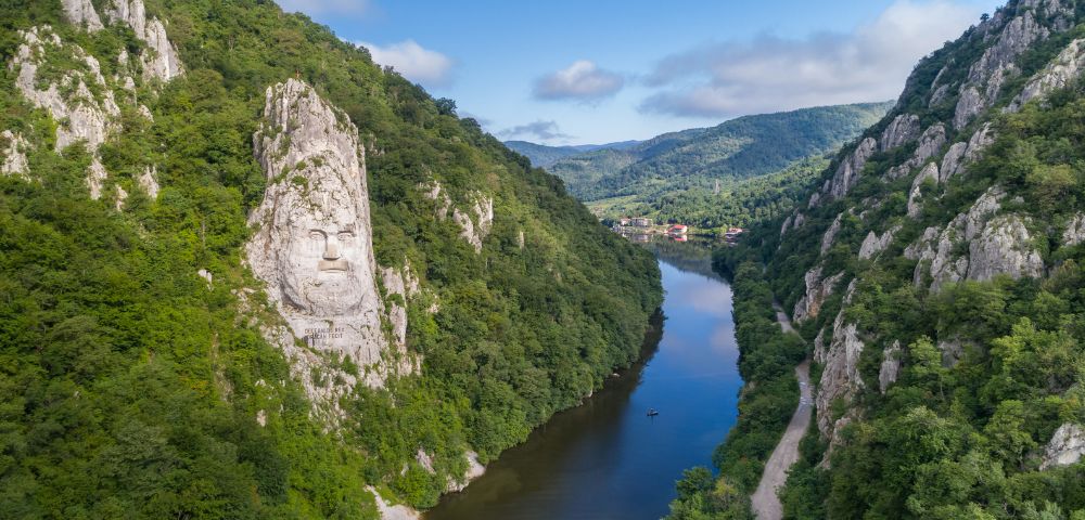 A serene river winds through a lush green canyon, flanked by steep hills. A large stone face is carved into one hill, under a clear blue sky.