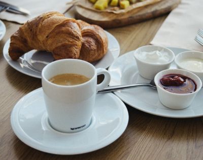 A breakfast table with a cup of coffee, two croissants on a plate, and small bowls of jam and cream. The scene conveys a cozy, inviting ambiance.