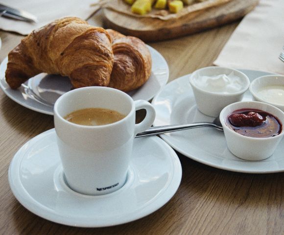 A breakfast table with a cup of coffee, two croissants on a plate, and small bowls of jam and cream. The scene conveys a cozy, inviting ambiance.