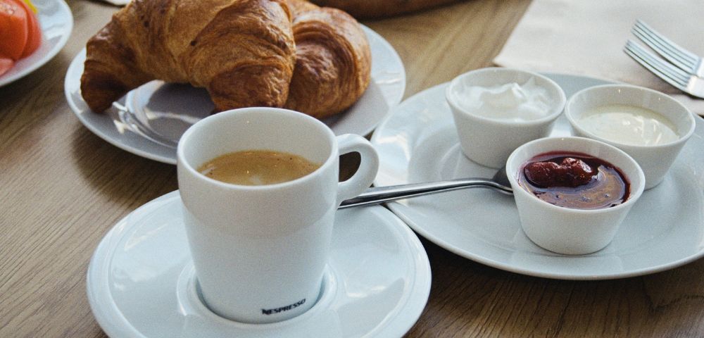 A breakfast table with a cup of coffee, two croissants on a plate, and small bowls of jam and cream. The scene conveys a cozy, inviting ambiance.