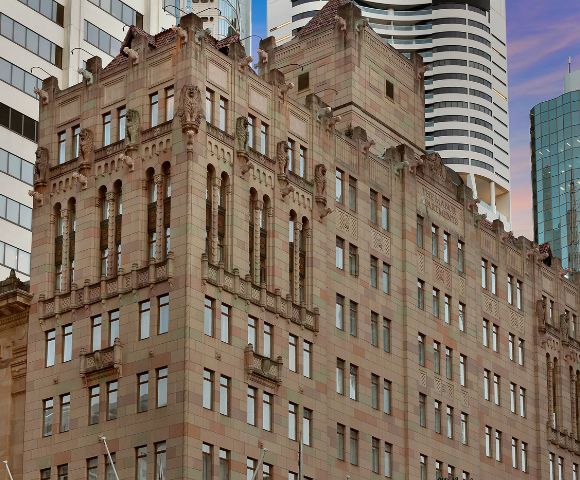Historic brownstone building with ornate architectural details, set against a backdrop of modern skyscrapers under a pink and blue sky at dusk.