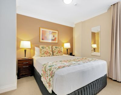 A cozy bedroom featuring a neatly made bed with floral pillows and a matching throw. Soft lighting from two lamps, warm tones, and a calming atmosphere.