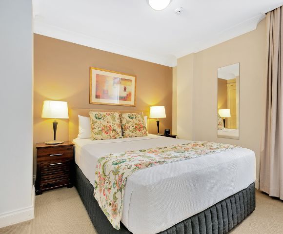 A cozy bedroom featuring a neatly made bed with floral pillows and a matching throw. Soft lighting from two lamps, warm tones, and a calming atmosphere.