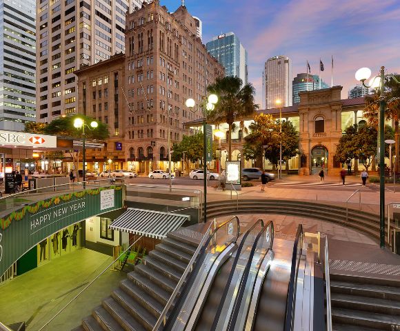 Urban scene at sunset with an illuminated square featuring historic and modern buildings. An escalator leads underground, surrounded by city lights.