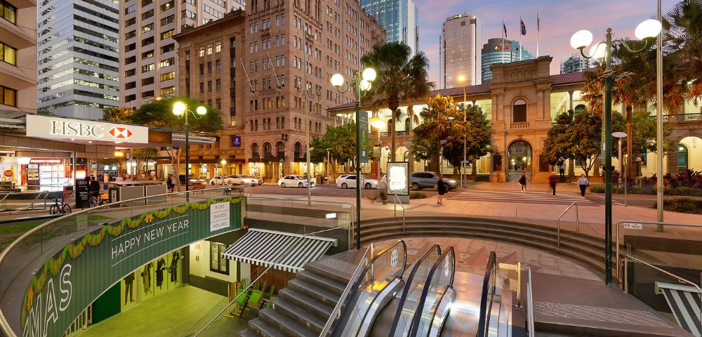 Urban scene at sunset with an illuminated square featuring historic and modern buildings. An escalator leads underground, surrounded by city lights.