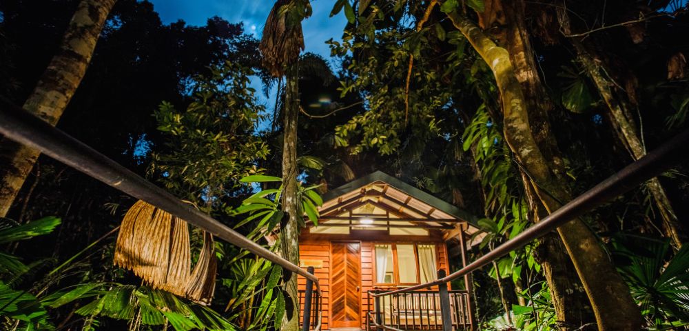 Wooden cabin nestled in a lush, tropical rainforest, viewed from a pathway at dusk. Warm light glows from inside, creating an inviting, tranquil atmosphere.