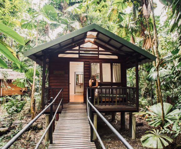 Wooden cabin in lush jungle, accessible via a wooden ramp. A person stands on the porch beneath a metal roof, surrounded by dense green foliage.