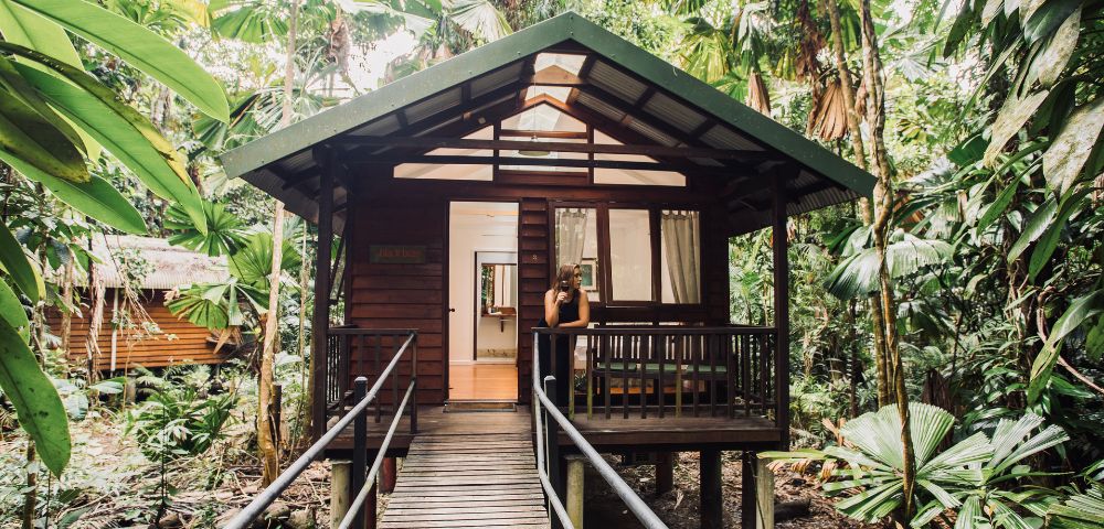 Wooden cabin in lush jungle, accessible via a wooden ramp. A person stands on the porch beneath a metal roof, surrounded by dense green foliage.