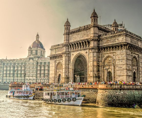 Iconic Gateway of India with ornate archways, flanked by the Taj Mahal Palace hotel. Boats on the water suggest a bustling, historic waterfront scene.