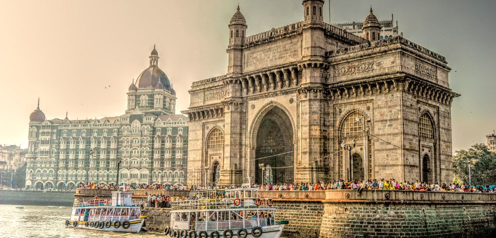 Iconic Gateway of India with ornate archways, flanked by the Taj Mahal Palace hotel. Boats on the water suggest a bustling, historic waterfront scene.