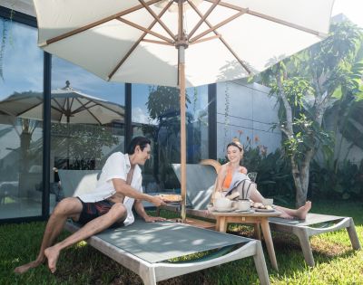 A man and woman relax on sun loungers under a large umbrella in a lush garden, sharing refreshments. The scene is calm and sunny, evoking a sense of leisure.